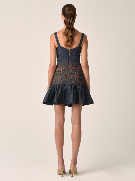 A woman with long hair in a ponytail stands facing away, wearing a sleeveless dress featuring the Lucile Skirt Chambray Embroidery with a fitted waist that accentuates her feminine silhouette. She completes her look with sleek beige heels.