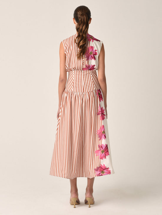 A woman is stylishly dressed in the Jocelyn Dress in Magenta Floral Brushstroke Stripes, featuring vertical orange accents, and paired with open-toed shoes for a charming daytime ensemble.