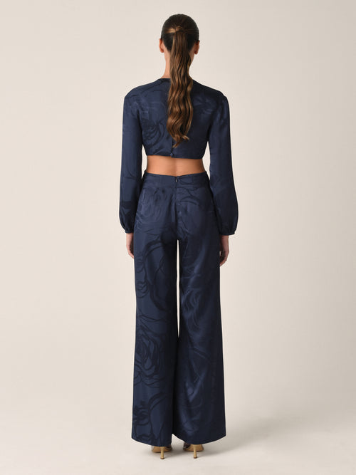 The Joely Jumpsuit Navy Jacquard, a black long-sleeve piece from the Resort 2023 collection, features a twisted front design, cutout at the waist, and wide-leg trousers.