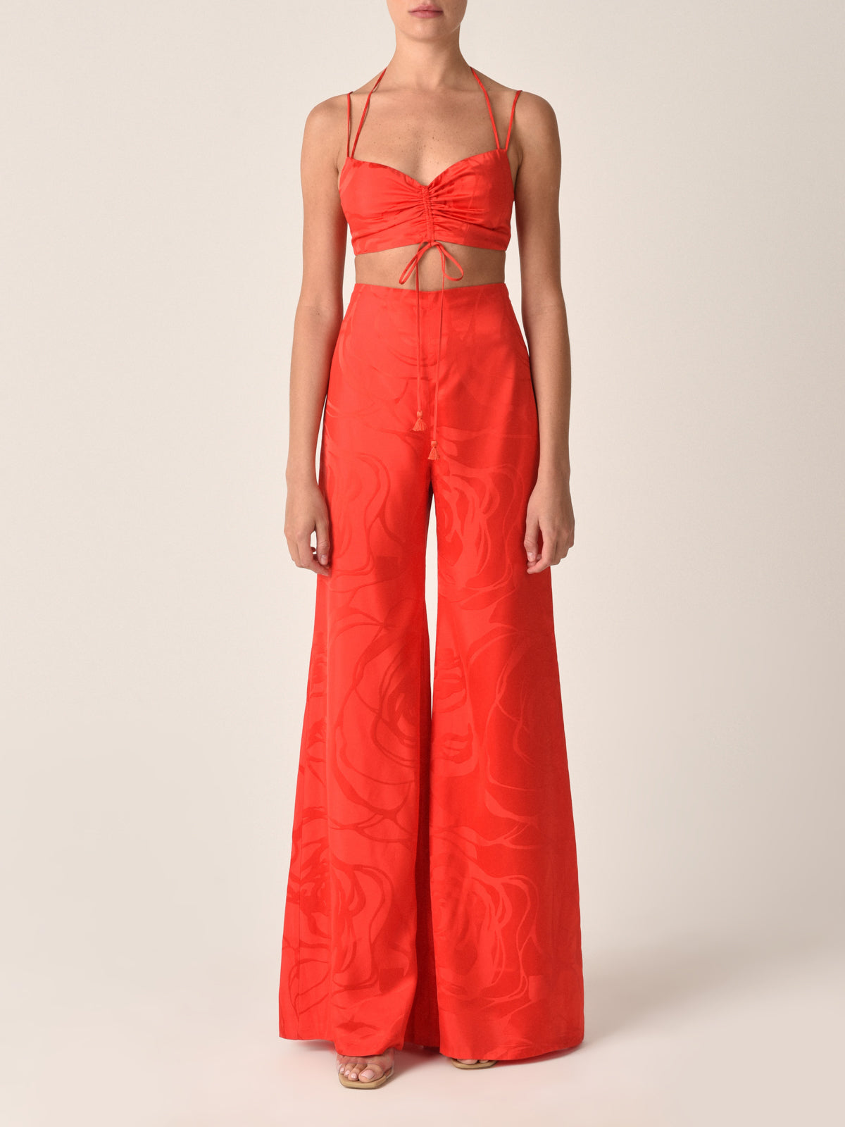 The Kayla Top Crimson Jacquard is a cropped camisole with thin straps and a halter neck tie, featuring ruching detail at the front and long tassel ties hanging down.