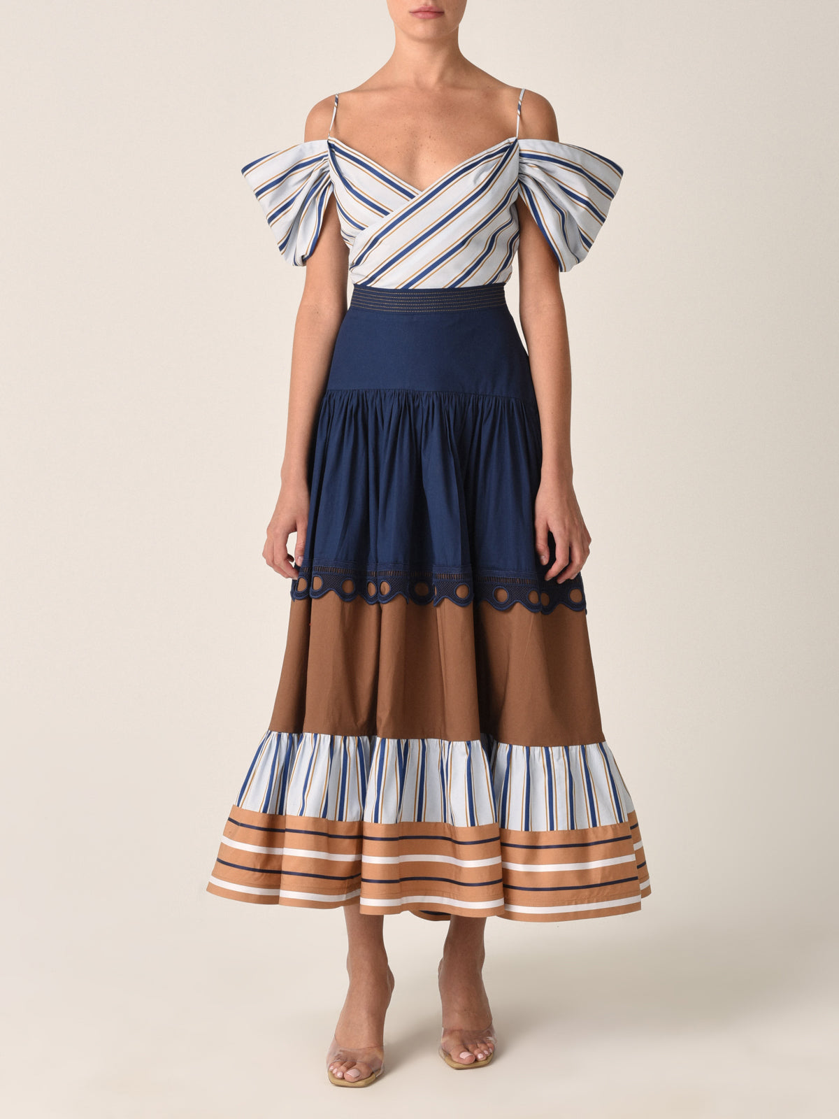 The Justine Skirt Camel Navy Stripes is a long, tiered midi skirt featuring a navy blue top section, a brown middle section adorned with cotton guipure lace detail, and a striped bottom section in shades of blue, brown, and white.