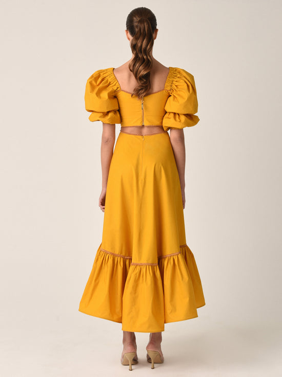 Introducing the Peggie Dress Marigold, a stunning Silvia Tcherassi dress in mustard yellow, featuring puffed sleeves, a fitted bodice, side cutouts, and a ruffled hem—an essential piece from Resort 2023.