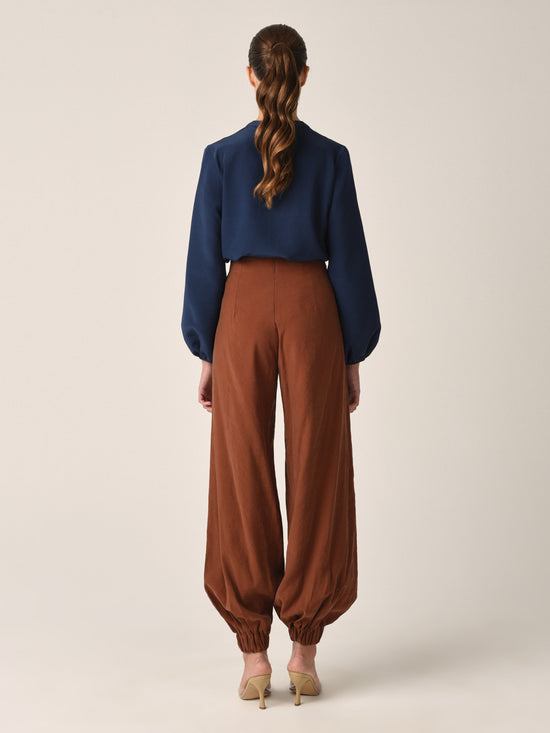 The Raimondo Pant Cacao, featuring a high-waisted design with a twisted front detail and gathered cuffs, pairs perfectly with our honey blouse crafted from luxurious vegan silk.