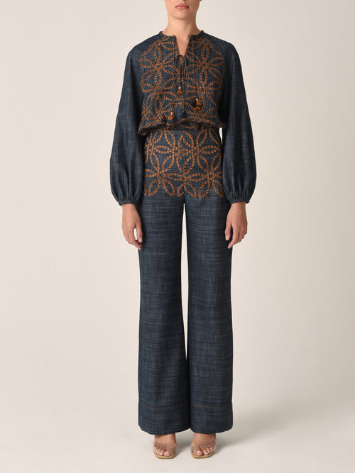 A dark blue long-sleeved Vigolene Blouse Chambray Embroidery featuring an intricate geometric pattern in orange. The high-fashion sleeves add an elegant touch, while a tie at the neckline with tassels completes this chic look.