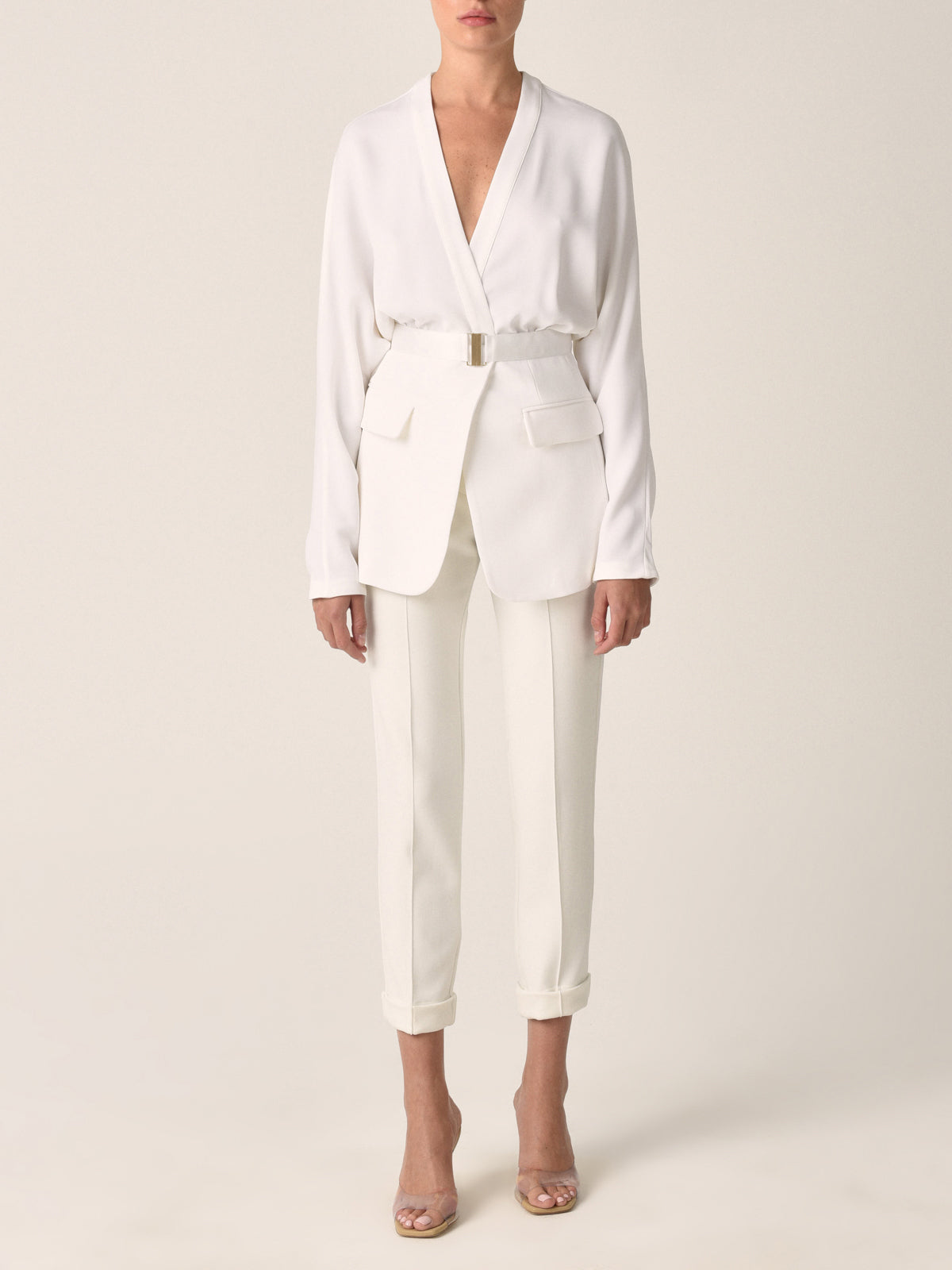 A white belted blazer with long sleeves, two front pockets, and a V-neck collar photographed against a plain background exudes modern elegance. This seasonless sleek white viscose piece, known as the Vogogna Jacket White, is a timeless addition to any wardrobe.