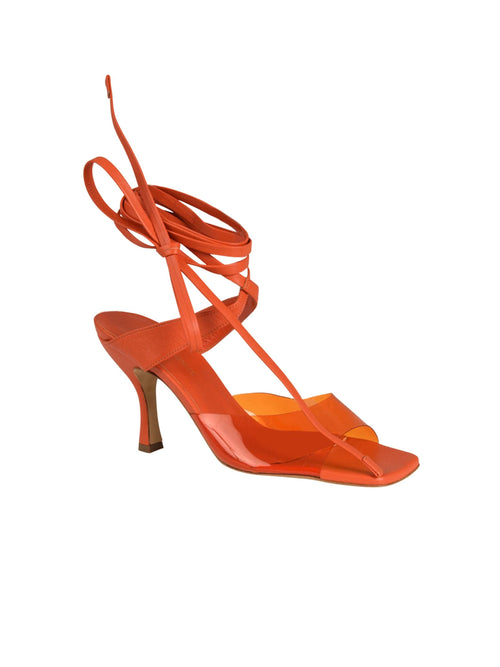 The Marlene Heels Orange feature a bold orange strappy sandal crafted from luxurious Italian calf leather, complete with a mid-heel and sophisticated ankle tie design, elegantly visible from the side.