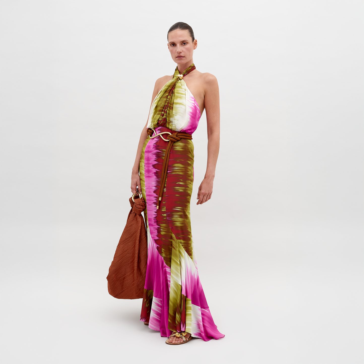 A person dressed in a Raisa Dress Violeta Lime Tie Dye with pink, green, and white patterns, holding a large brown bag, posing against a plain background. Pre-order by November 15th 2024 to ensure the perfect ship-by date.