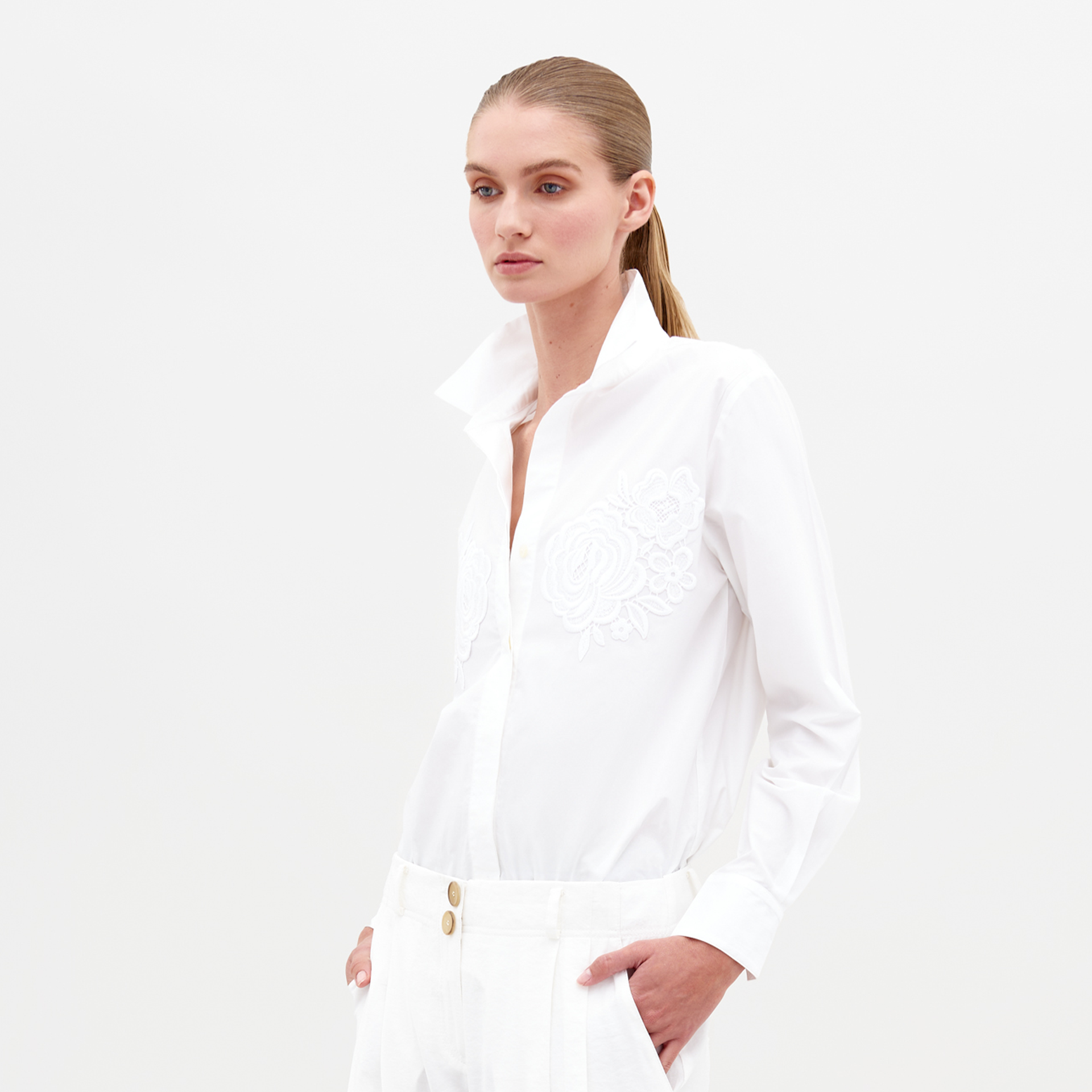 Sporting the Raizel Blouse White, a person with long hair stands against a plain background with hands in pockets, dressed in a white embroidered button-up shirt with the collar turned up and coordinating white pants. They are ready for the pre-order shoot scheduled for February 15th, 2025.