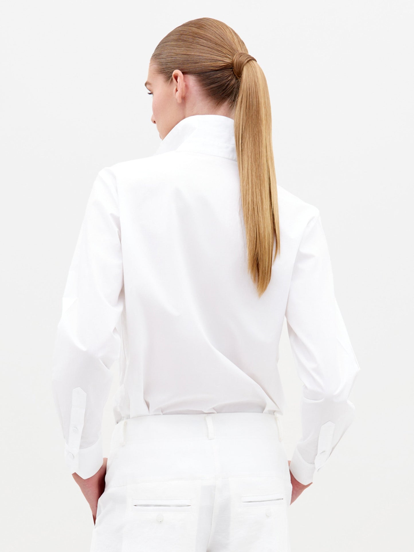 Sporting the Raizel Blouse White, a person with long hair stands against a plain background with hands in pockets, dressed in a white embroidered button-up shirt with the collar turned up and coordinating white pants. They are ready for the pre-order shoot scheduled for February 15th, 2025.