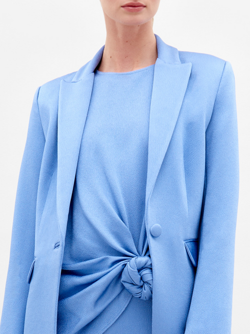 A person in a blue Rebeca Jacket with a single-button closure and sandals, holding a beige bag, stands against a plain white background, exuding the elegance of fine Italian textile.