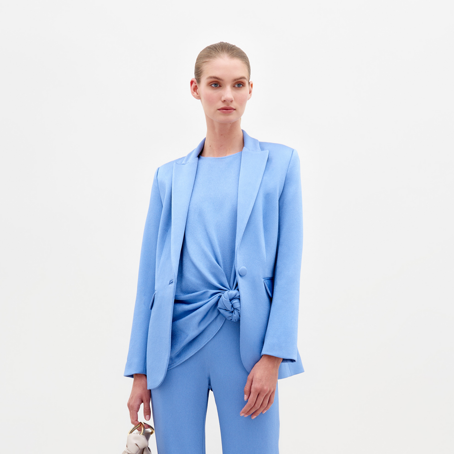 A person in a blue Rebeca Jacket with a single-button closure and sandals, holding a beige bag, stands against a plain white background, exuding the elegance of fine Italian textile.