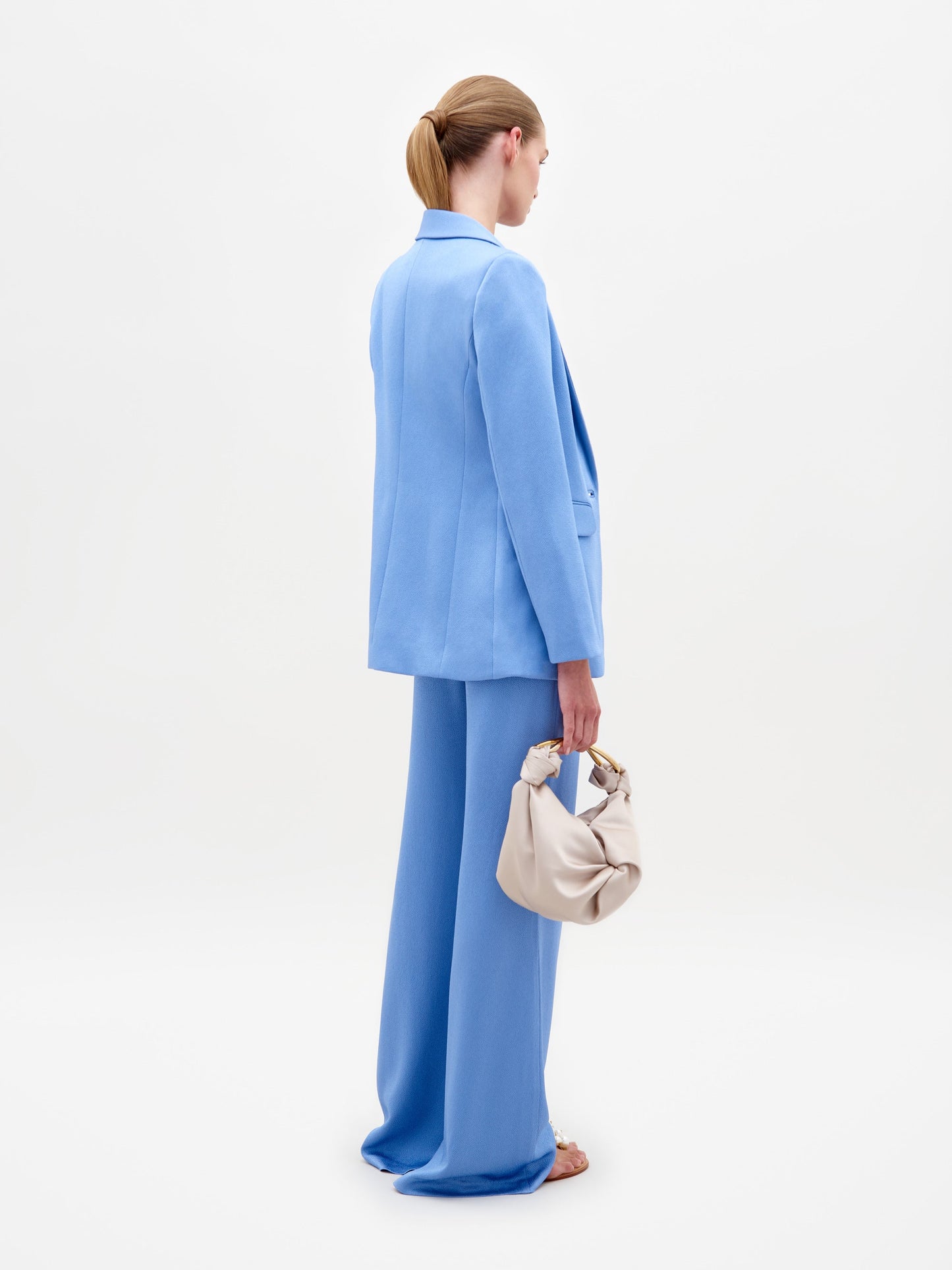 A person in a blue Rebeca Jacket with a single-button closure and sandals, holding a beige bag, stands against a plain white background, exuding the elegance of fine Italian textile.