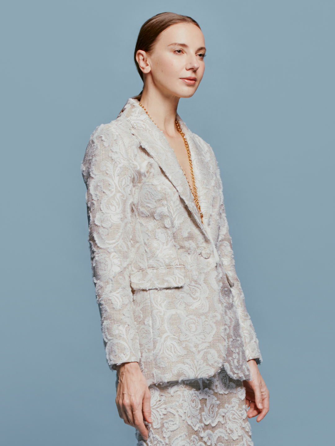 A person with slicked-back hair wears the Rebeca Jacket in Abstract White Floral and matching pants, looking to the side against a blue background. Mark your calendars for September 2nd, 2024; outfits like this will ship between August 19th and that date.