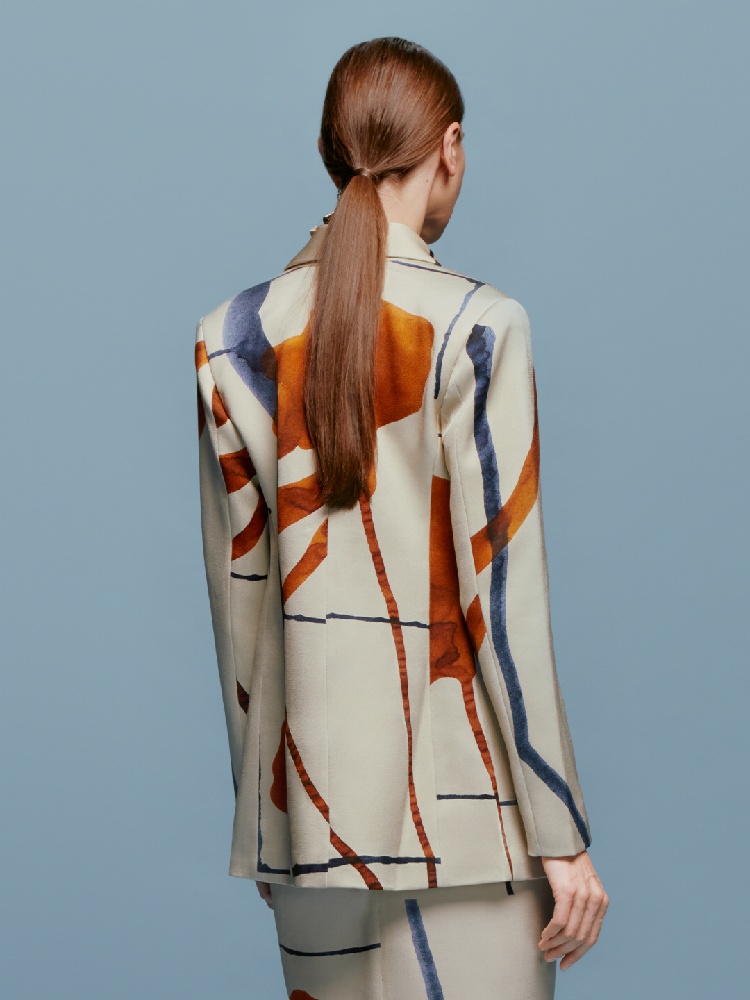 A woman with tied back hair stands against a blue background, wearing the Rebeca Jacket in Cobalt Copper Abstract with a matching dress, and large, abstract earrings, ready for her feature on September 2nd.