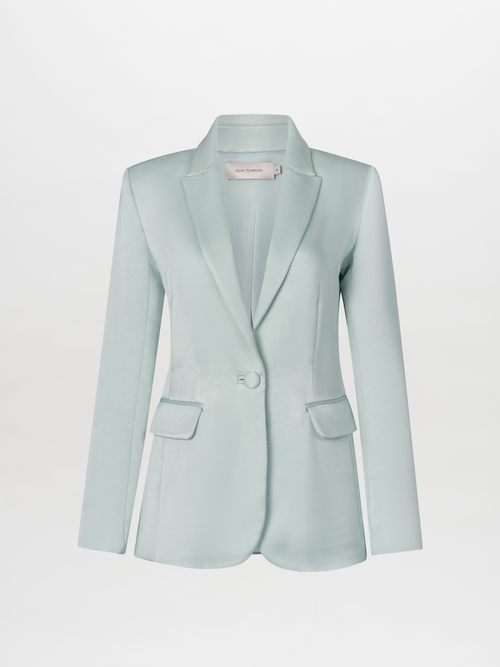 A woman in a turquoise Rebeca Jacket over a burgundy top stands against a light blue background. She has straight, pulled-back hair and an "S" charm with an attached trinket hanging from her jacket pocket, ready to make a statement on September 2nd.