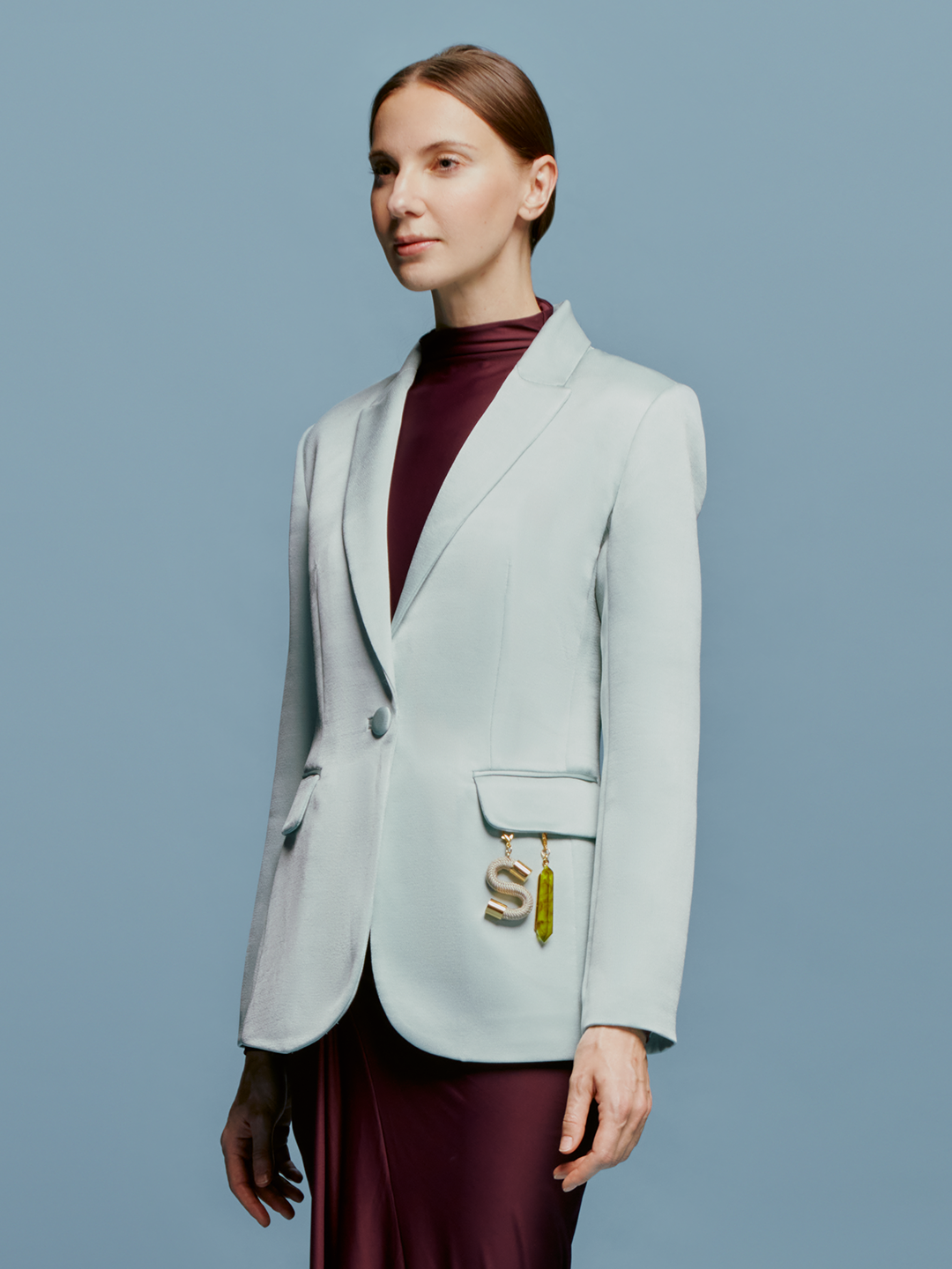 A woman in a turquoise Rebeca Jacket over a burgundy top stands against a light blue background. She has straight, pulled-back hair and an "S" charm with an attached trinket hanging from her jacket pocket, ready to make a statement on September 2nd.