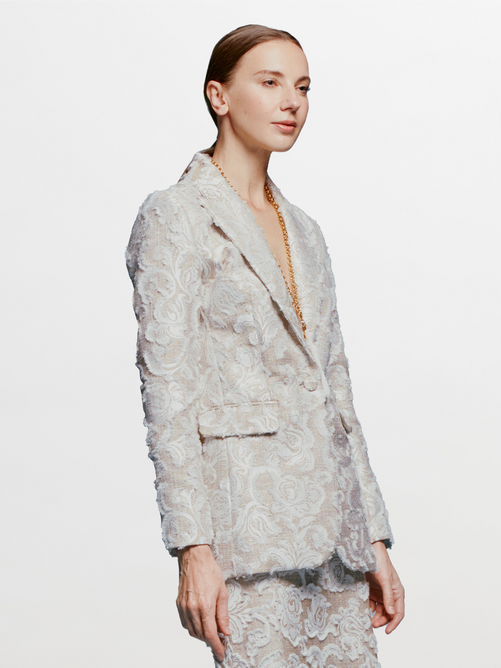 A person with slicked-back hair wears the Rebeca Jacket in Abstract White Floral and matching pants, looking to the side against a blue background. Mark your calendars for September 2nd, 2024; outfits like this will ship between August 19th and that date.