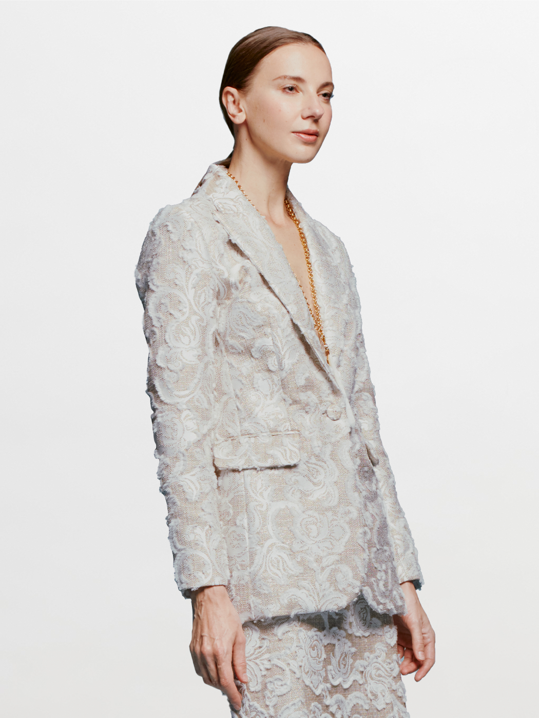 A person with slicked-back hair wears the Rebeca Jacket in Abstract White Floral and matching pants, looking to the side against a blue background. Mark your calendars for September 2nd, 2024; outfits like this will ship between August 19th and that date.