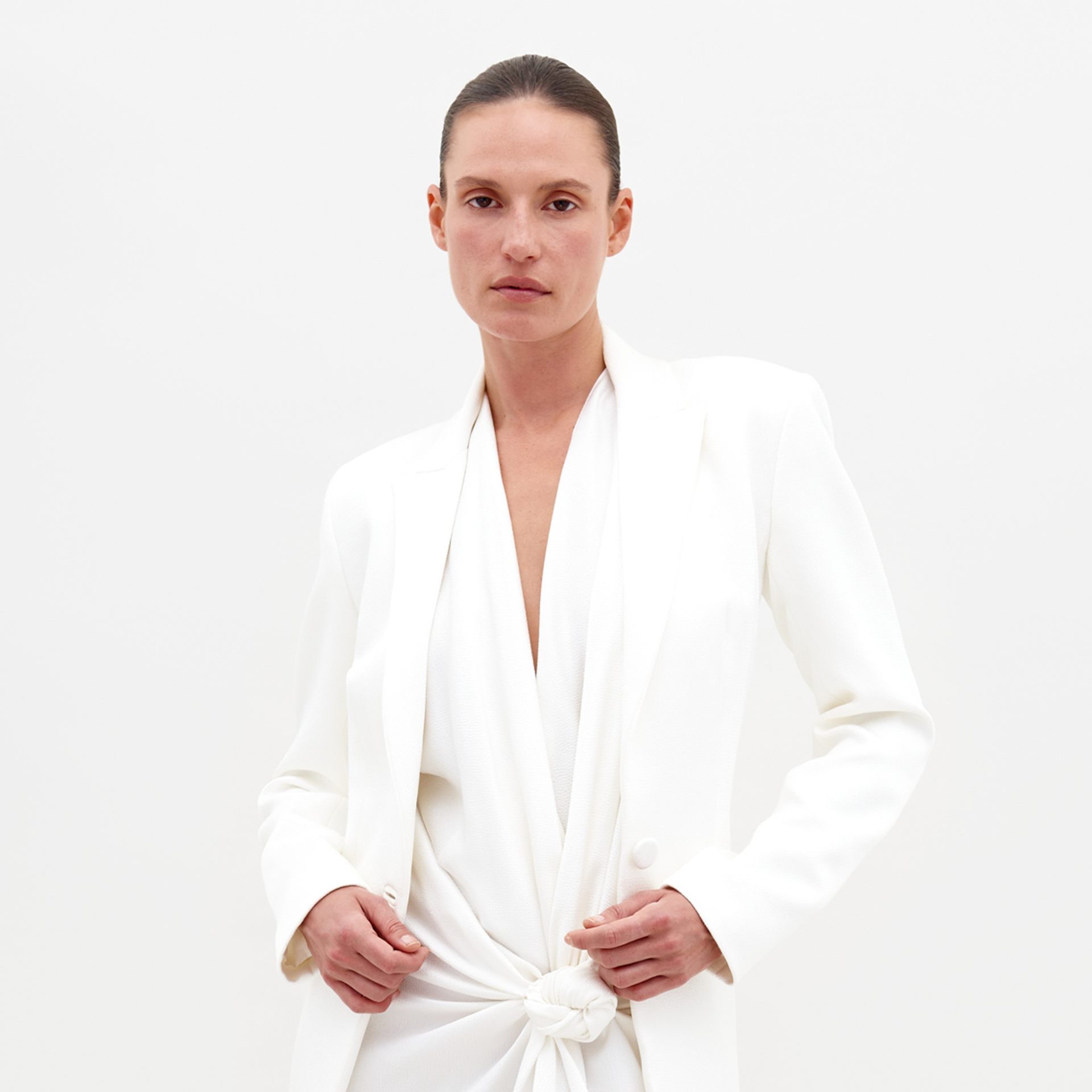 A person with a neutral expression is wearing the Rebeca Jacket White and white pants, standing against a plain background with arms crossed, available for pre-order starting February 15th, 2025.