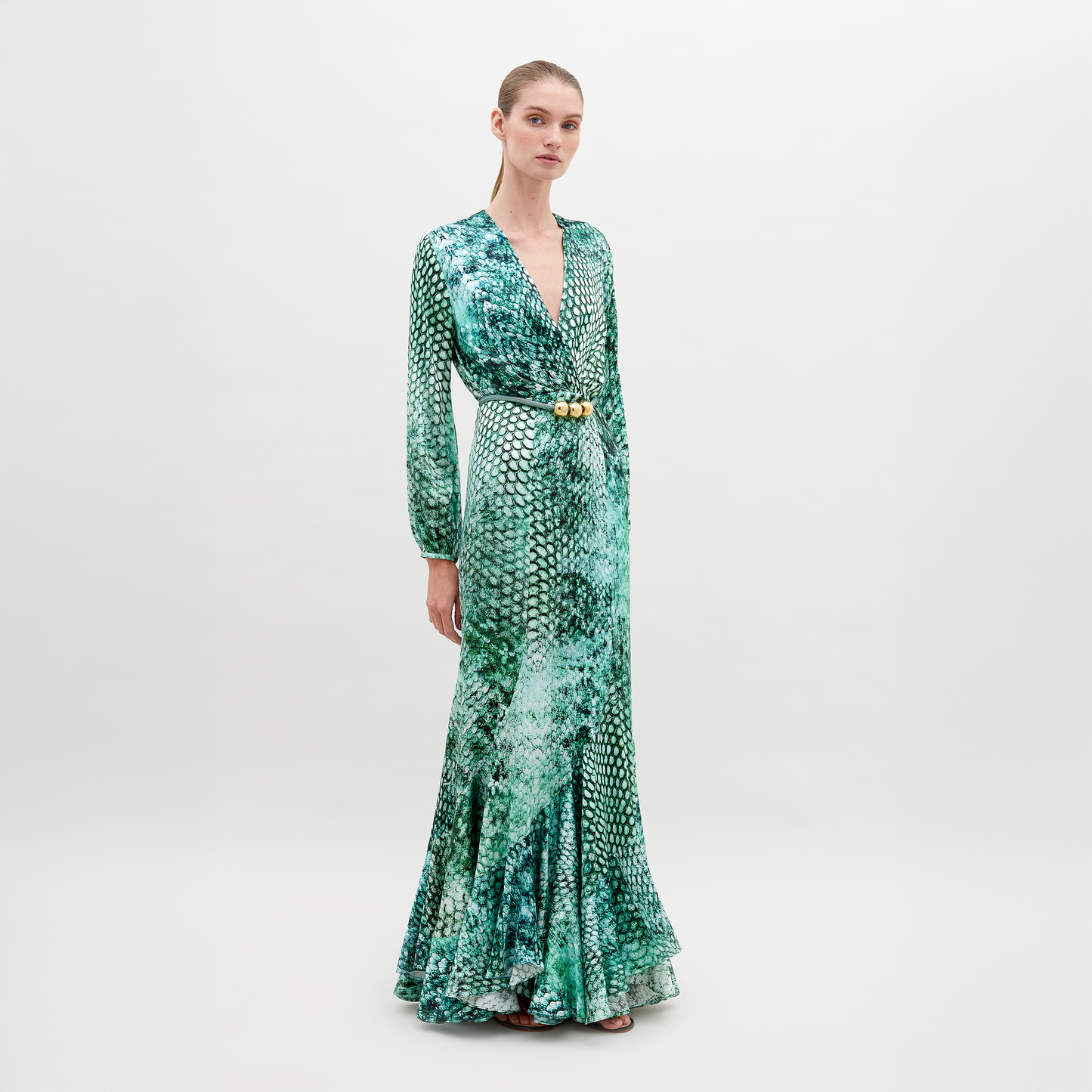 A person stands wearing the Remy Dress Emerald Animal Print, a green long-sleeve, full-length dress with a pattern resembling scales, featuring a V-neckline and a belt with ornamental details. Available for pre-order now, this stunning piece will ship by date November 15th, 2024. The background is plain white.