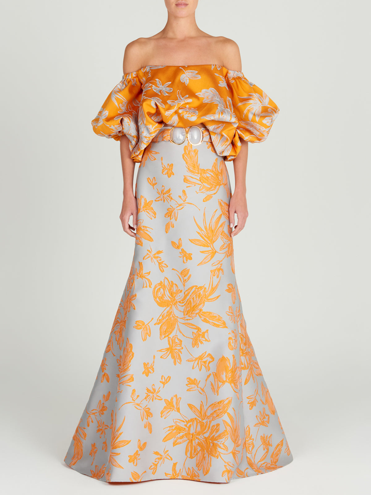 The Arezzo Blouse Apricot Silver showcases a charming orange off-the-shoulder design with a white floral pattern and voluminous sleeves, exuding polished elegance against a plain background.