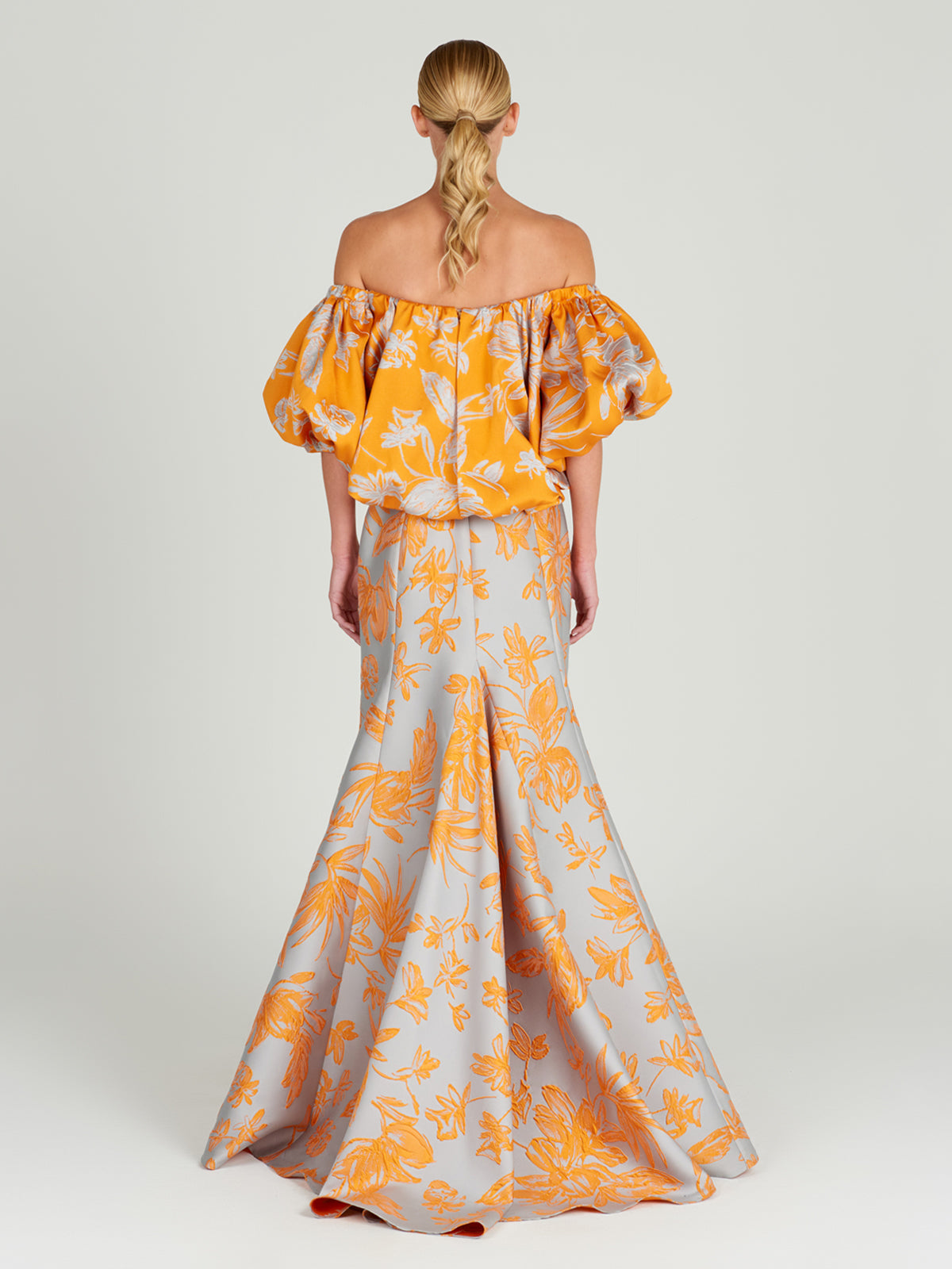 The Arezzo Blouse Apricot Silver showcases a charming orange off-the-shoulder design with a white floral pattern and voluminous sleeves, exuding polished elegance against a plain background.