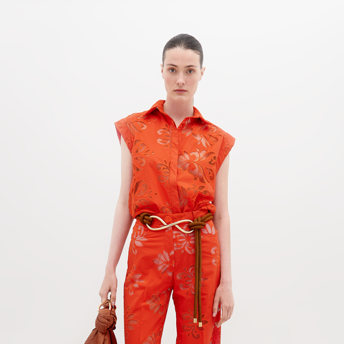 A person stands wearing a vibrant orange sleeveless top and matching pants with floral patterns, accessorized with a brown belt and holding a brown handbag. Pre-order the Rina Blouse Rouge Embroidery now to ensure it ships by the date of November 15th, 2024.