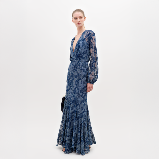 A person in a long, blue lace dress with a deep V-neckline and long sleeves stands against a plain white background, holding a black clutch—the Robin Dress Indigo Lace, available for pre-order, set to ship by November 15th, 2024.