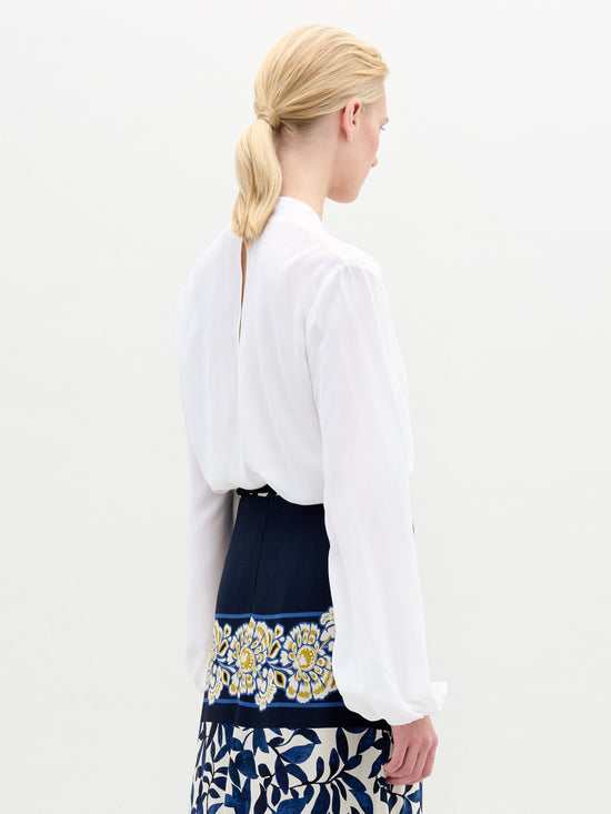 Individual wearing the Rocio Blouse, a white deep V-neck top, paired with a black and gold belt over a skirt adorned with floral patterns against a plain backdrop.