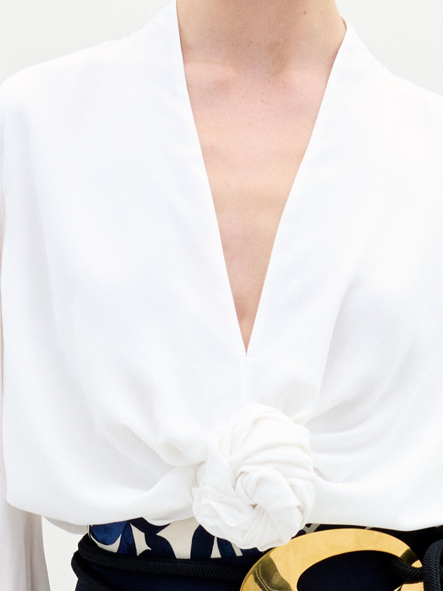 Individual wearing the Rocio Blouse, a white deep V-neck top, paired with a black and gold belt over a skirt adorned with floral patterns against a plain backdrop.