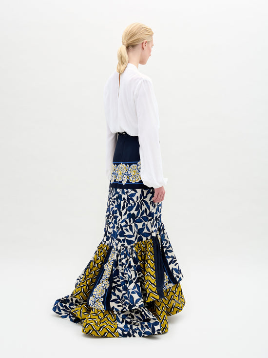 A model wears a white blouse paired with the Elliot Skirt, featuring a long design with a mix of blue, white, and yellow patterns.