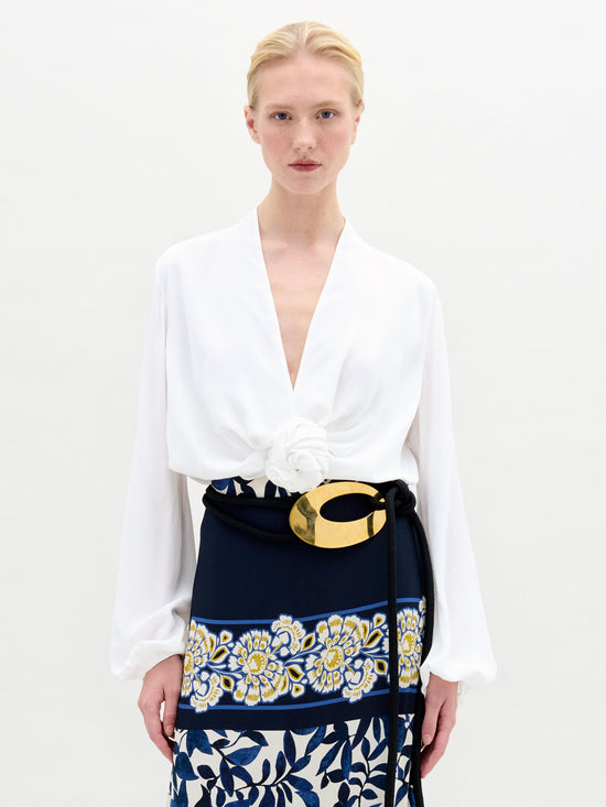 Individual wearing the Rocio Blouse, a white deep V-neck top, paired with a black and gold belt over a skirt adorned with floral patterns against a plain backdrop.