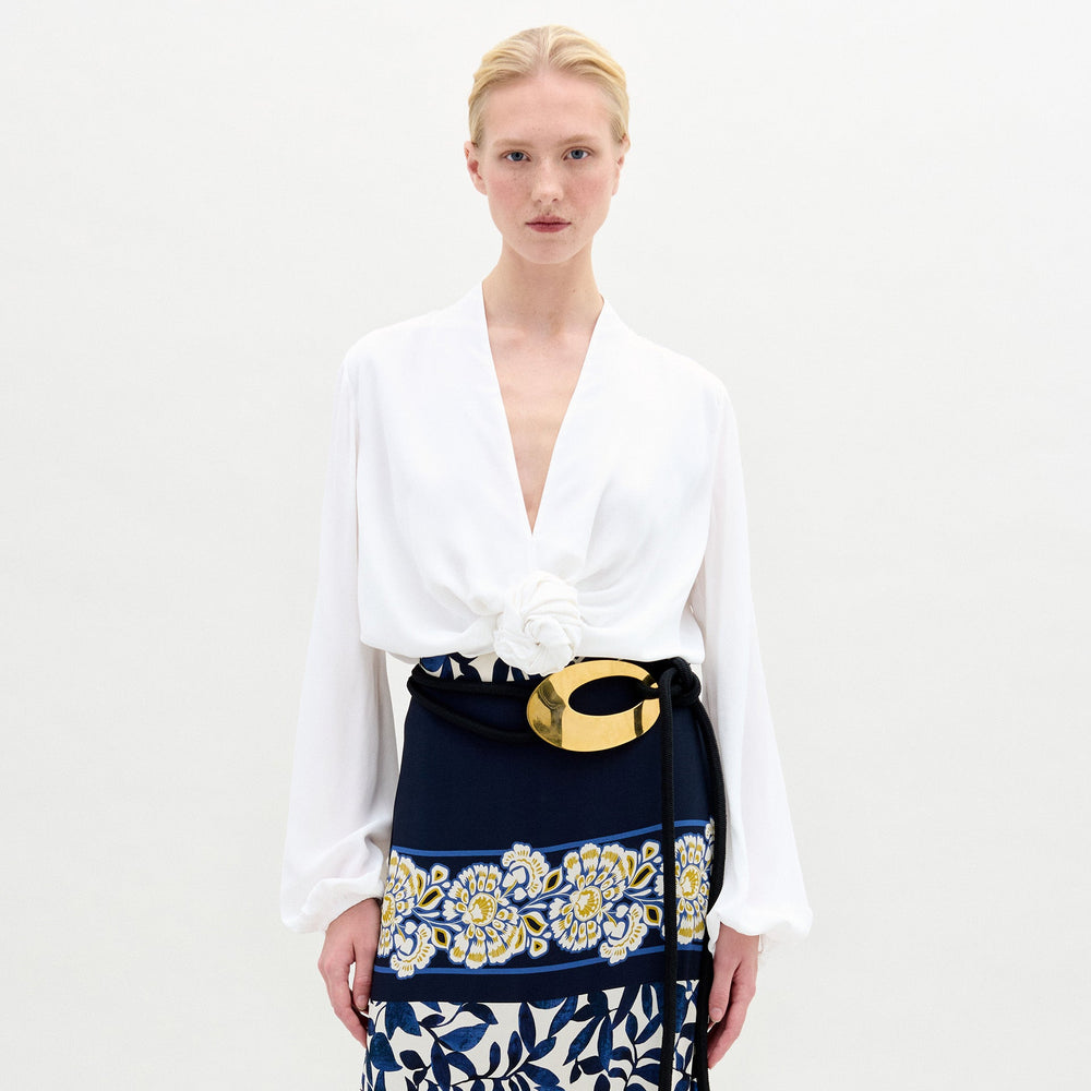 Model wearing the Rocio Blouse—a white, collarless V-neck top—paired with a floral belt featuring a gold buckle and a navy Italian fabric skirt with yellow and white floral prints, standing against a plain background.