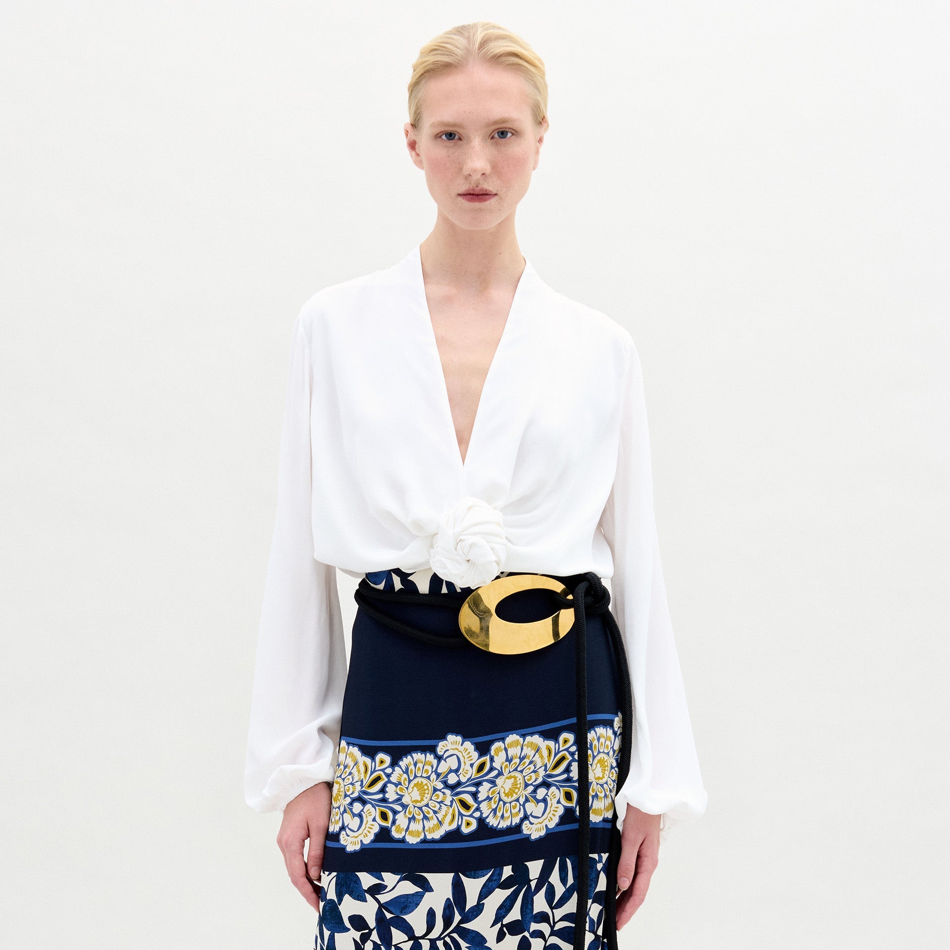 Model wearing the Rocio Blouse—a white, collarless V-neck top—paired with a floral belt featuring a gold buckle and a navy Italian fabric skirt with yellow and white floral prints, standing against a plain background.