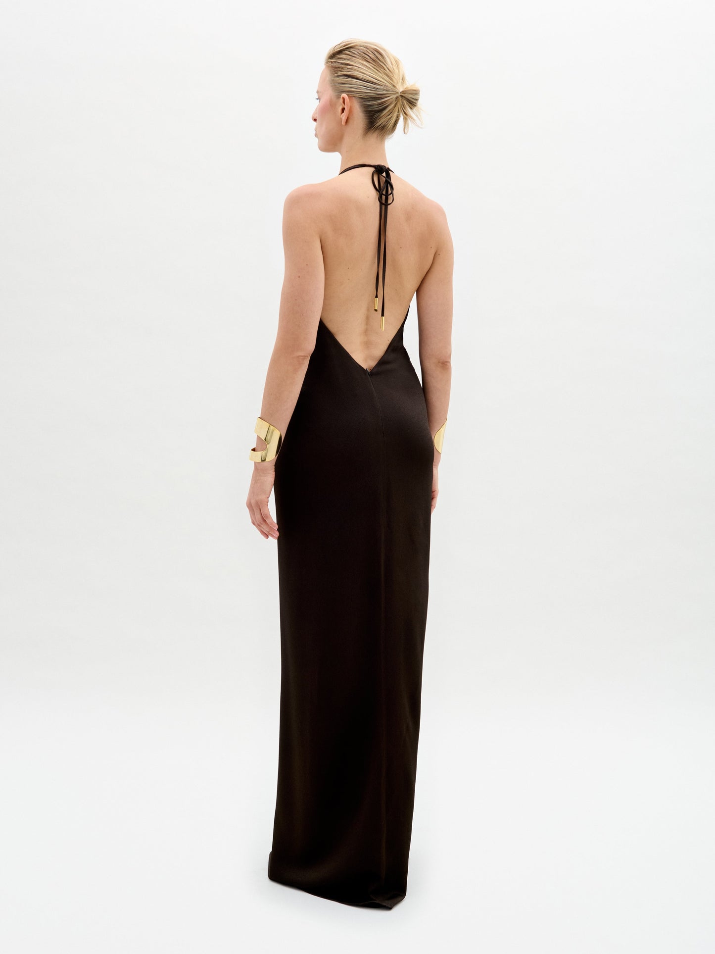 A model poses against a plain backdrop in the Rowan Dress, a sleek black halter gown featuring a deep neckline and gold belt. She completes the look with matching gold bracelets and shoes.