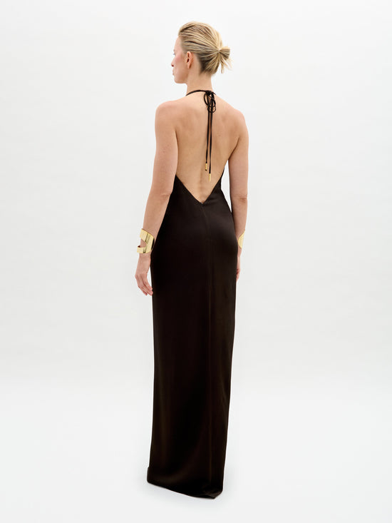 A model poses against a plain backdrop in the Rowan Dress, a sleek black halter gown featuring a deep neckline and gold belt. She completes the look with matching gold bracelets and shoes.