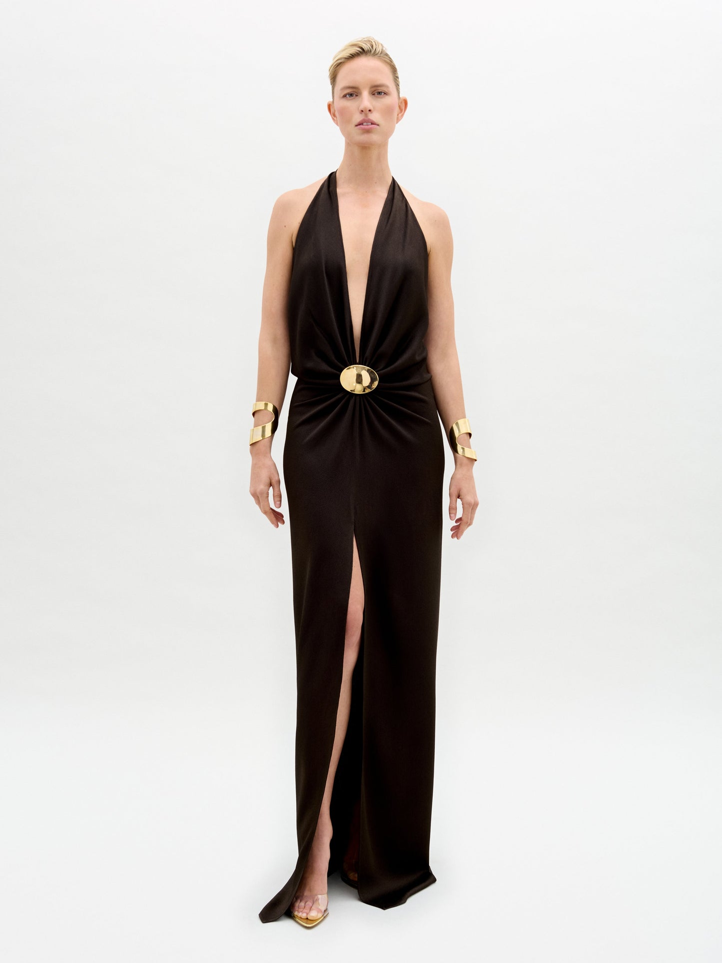 A model poses against a plain backdrop in the Rowan Dress, a sleek black halter gown featuring a deep neckline and gold belt. She completes the look with matching gold bracelets and shoes.