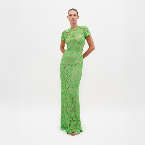 A person stands against a plain white background, wearing the Roxana Dress Green Guipure—a long, fitted lace dress with short sleeves. Hands are clasped behind the back. This elegant dress is available for pre-order now, with a scheduled ship date of February 15th, 2025.