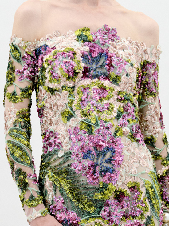 A person stands against a plain background wearing the Rafaella Dress Botanical Sequins, a floor-length, off-the-shoulder floral gown with long sleeves and adorned with a vibrant pattern of flowers and greenery. This elegant piece is available for pre-order now, with a ship-by date set for February 2025.