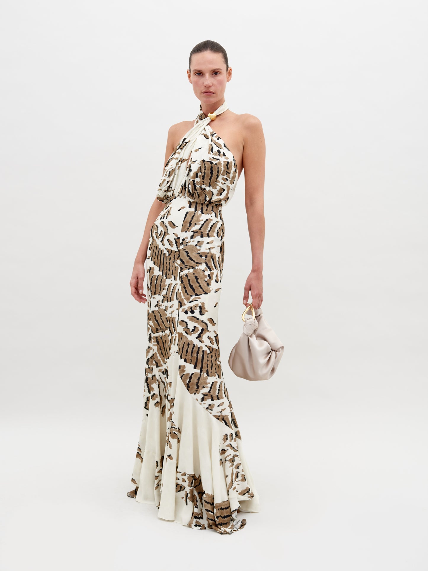 A woman stands in a Raisa Dress Beige Abstract Animal Print with a halter neckline, holding a small, light-colored handbag. She has a neutral expression and stands against a plain white background. This chic ensemble is available for pre-order, with a ship by date of November 15th, 2024.