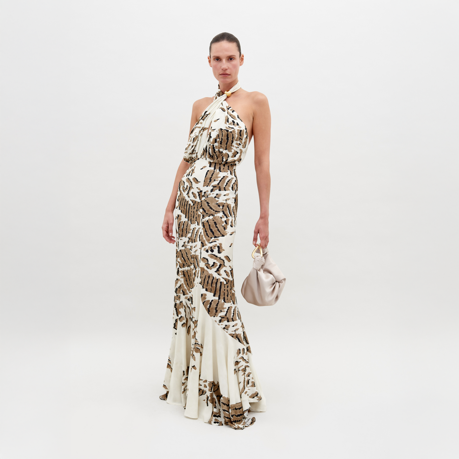 A woman stands in a Raisa Dress Beige Abstract Animal Print with a halter neckline, holding a small, light-colored handbag. She has a neutral expression and stands against a plain white background. This chic ensemble is available for pre-order, with a ship by date of November 15th, 2024.