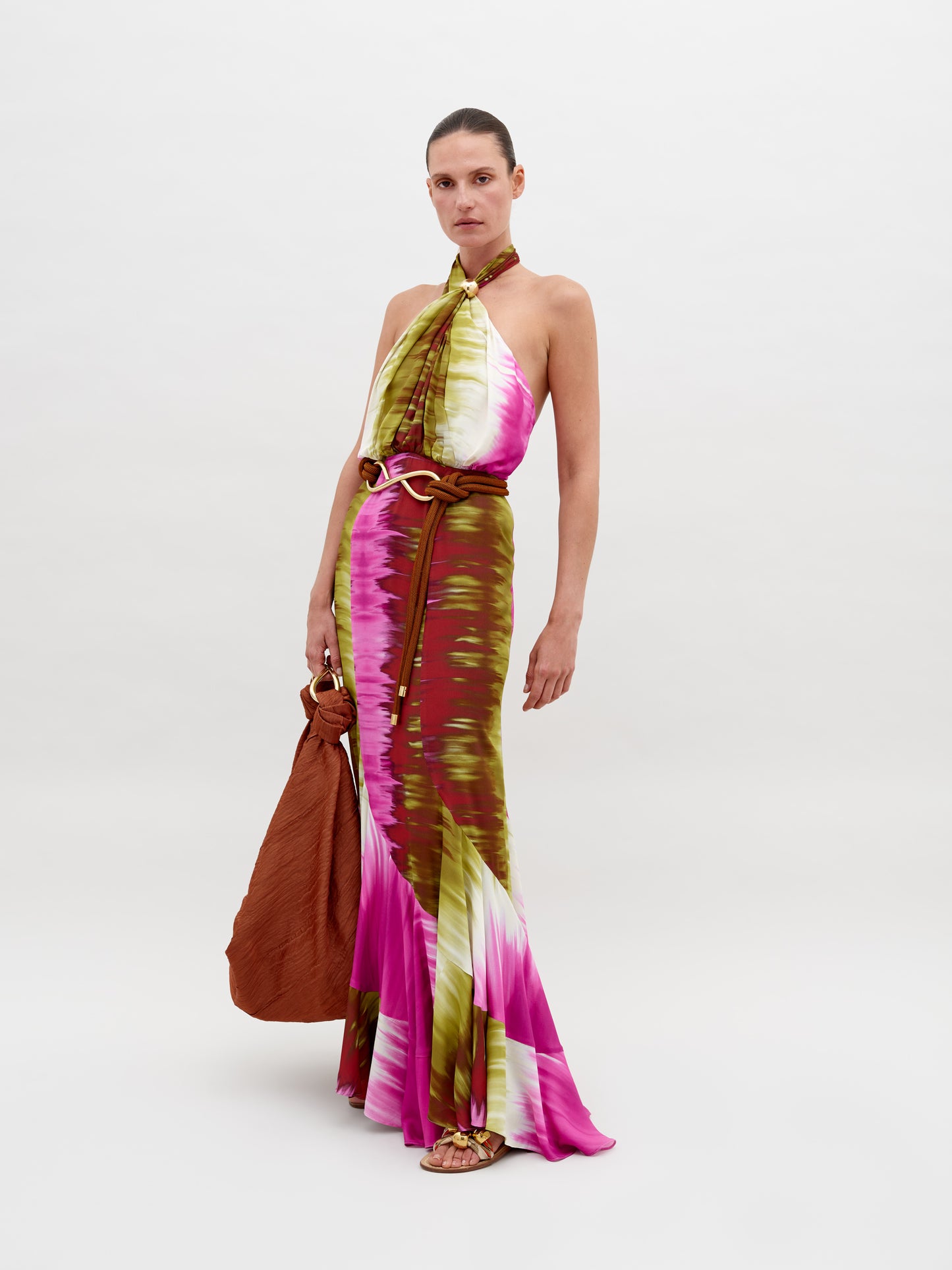 A person dressed in a Raisa Dress Violeta Lime Tie Dye with pink, green, and white patterns, holding a large brown bag, posing against a plain background. Pre-order by November 15th 2024 to ensure the perfect ship-by date.