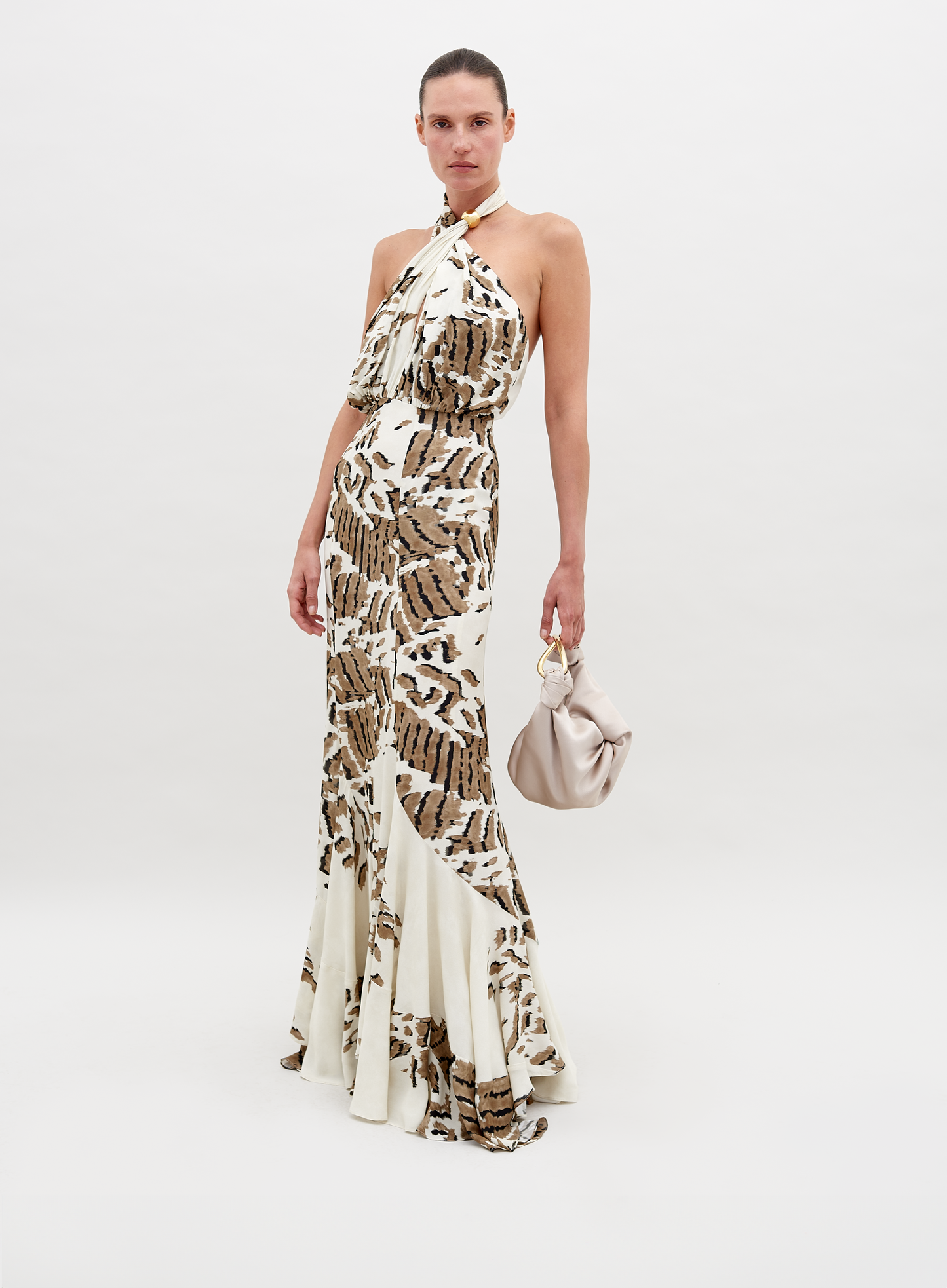 A woman stands in a Raisa Dress Beige Abstract Animal Print with a halter neckline, holding a small, light-colored handbag. She has a neutral expression and stands against a plain white background. This chic ensemble is available for pre-order, with a ship by date of November 15th, 2024.