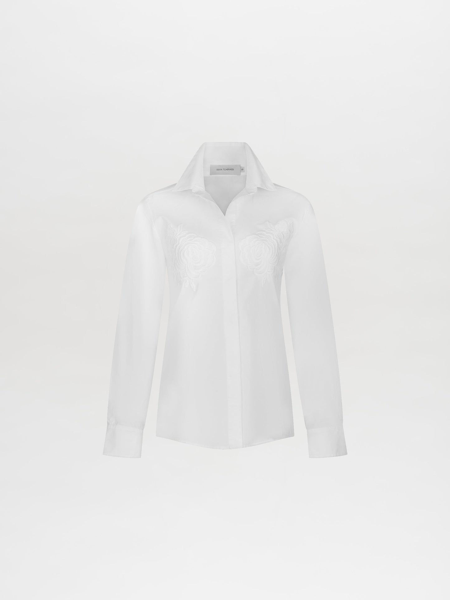 Sporting the Raizel Blouse White, a person with long hair stands against a plain background with hands in pockets, dressed in a white embroidered button-up shirt with the collar turned up and coordinating white pants. They are ready for the pre-order shoot scheduled for February 15th, 2025.