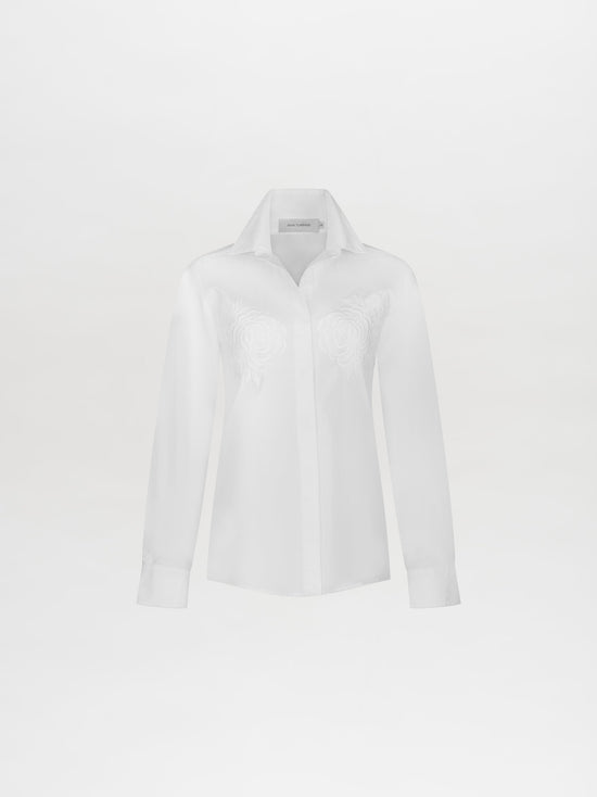 Sporting the Raizel Blouse White, a person with long hair stands against a plain background with hands in pockets, dressed in a white embroidered button-up shirt with the collar turned up and coordinating white pants. They are ready for the pre-order shoot scheduled for February 15th, 2025.