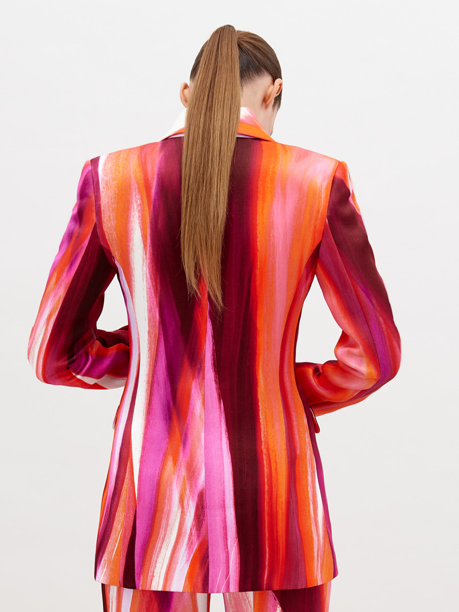 Woman wearing a vibrant, multi-colored striped Rebeca Jacket Magenta Abstract Waves with a deep neckline, accessorized with a chunky gold choker necklace, standing against a plain white background. Pre-order now and have it ship by date of November 15th, 2024.