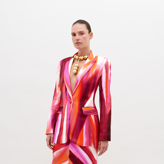 Woman wearing a vibrant, multi-colored striped Rebeca Jacket Magenta Abstract Waves with a deep neckline, accessorized with a chunky gold choker necklace, standing against a plain white background. Pre-order now and have it ship by date of November 15th, 2024.