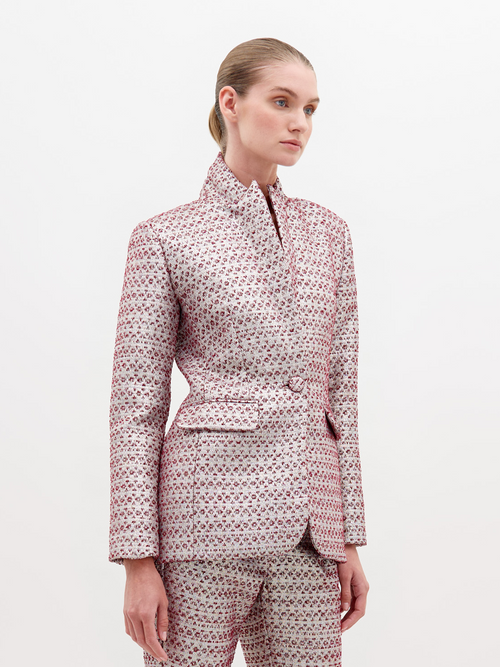 Woman in a Rebeca Jacket Metallic Burgundy and trousers stands against a plain white background, looking slightly to the right with a neutral expression. Pre-order now to secure this look; available exclusively starting November 15th, 2024.