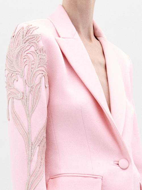 Profile view of a person with long hair tied back, wearing the Rebeca Jacket Pink, a light pink blazer adorned with intricate embroidery on the shoulder; pre-order now for delivery by February 1st, 2025.