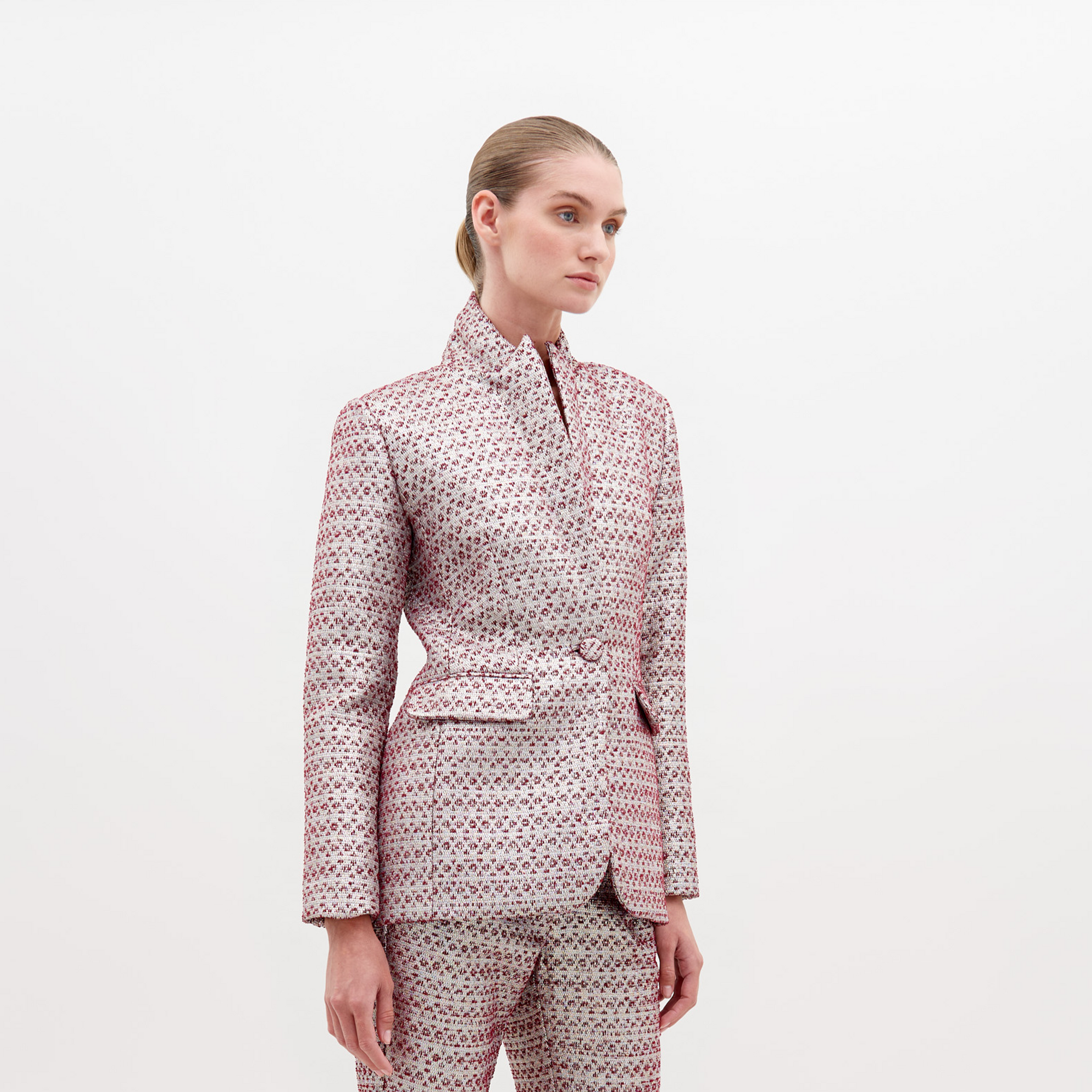 Woman in a Rebeca Jacket Metallic Burgundy and trousers stands against a plain white background, looking slightly to the right with a neutral expression. Pre-order now to secure this look; available exclusively starting November 15th, 2024.